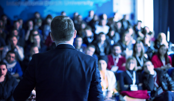 Audience seated facing a stage at a conference, seen from behind under blue lighting.