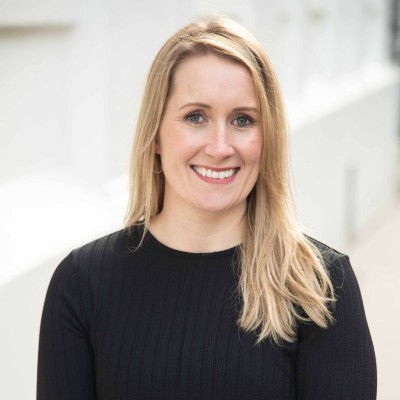 Professional headshot of a smiling blonde woman in a black top against a softly blurred light background.