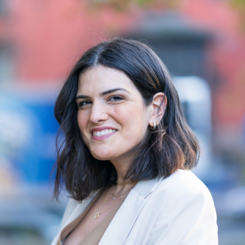 Smiling woman with shoulder-length dark hair wearing a light blazer, photographed outdoors with a blurred background.