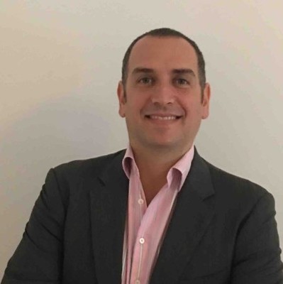 Headshot of a smiling man wearing a dark blazer and light pink collared shirt, standing against a plain light-colored wall.