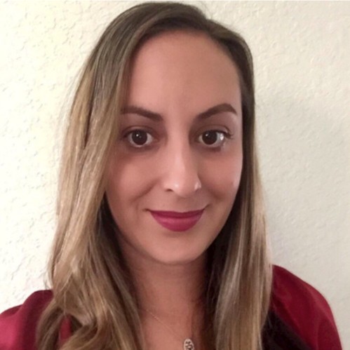 Smiling woman with long highlighted brown hair, wearing a burgundy top and pink lipstick, standing against a light textured wall.