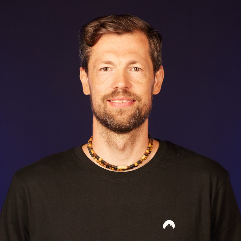 Smiling man with short brown hair and a beard, wearing a black shirt and a beaded necklace against a dark blue background.