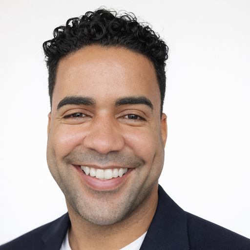 Headshot of a smiling man with short curly hair, wearing a dark blazer against a white background.