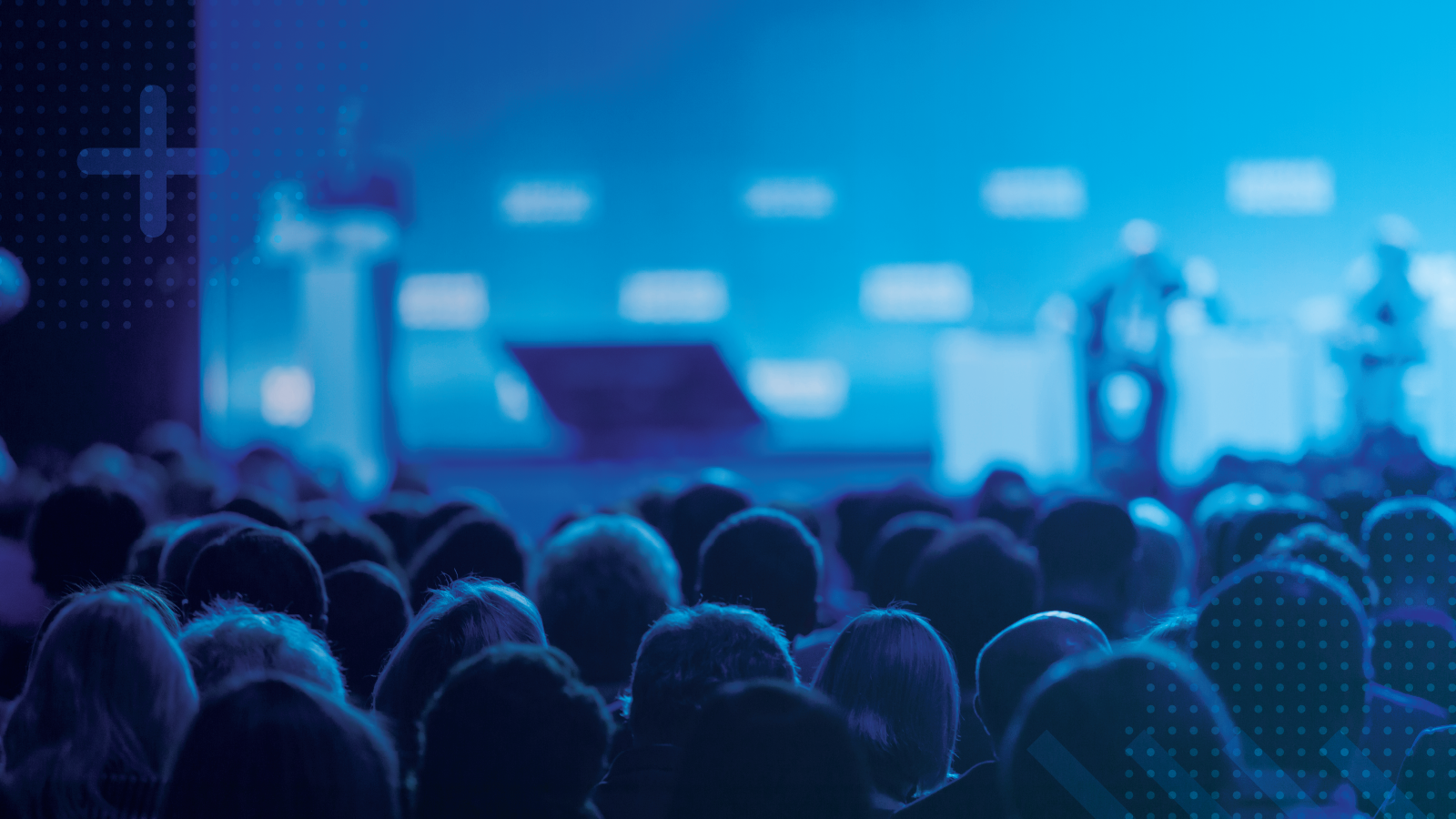 Audience seated in a dark auditorium watching a brightly lit blue stage with speakers and podiums in the background, suggesting a professional conference or industry event.