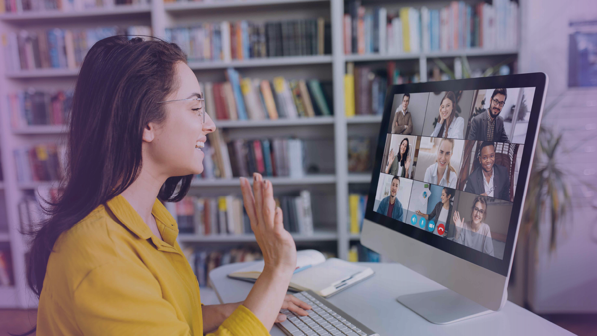Woman in a yellow shirt waves at her computer screen while participating in a virtual meeting with multiple people displayed in a video call grid.