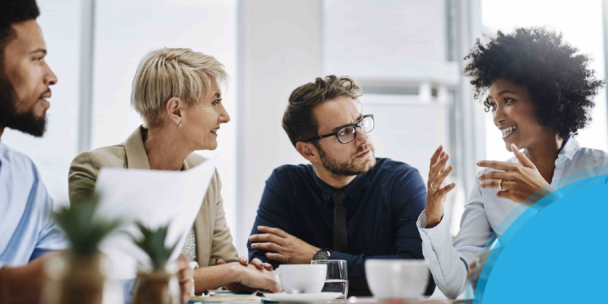 Four professionals engaged in a group discussion around a table, with a blue graphic overlay in the lower-right corner.