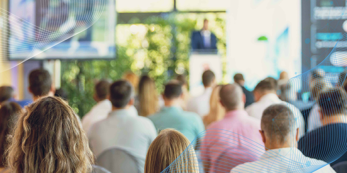 Audience seated at a conference presentation, viewed from behind, with abstract blue wave graphics.