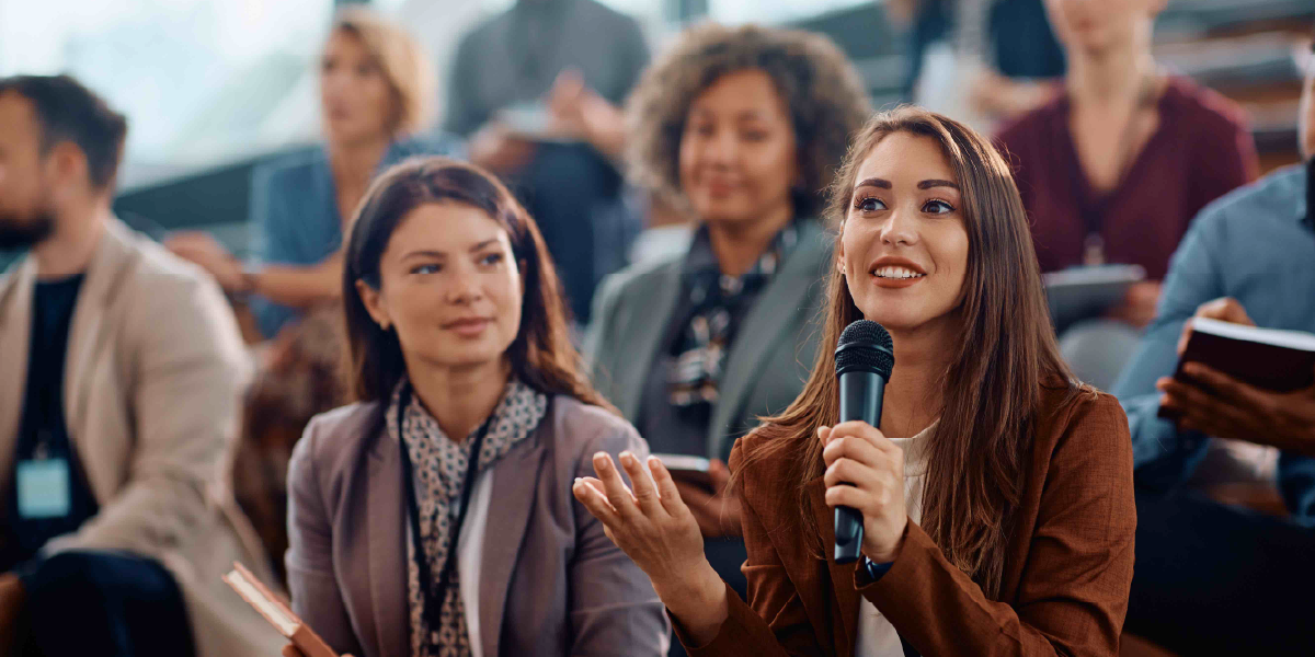 Woman speaking into a microphone during a conference Q&A as other attendees listen.