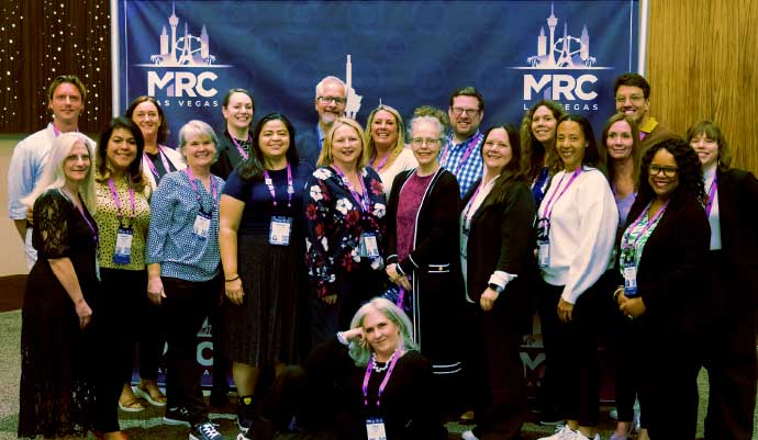  Group photo of MRC staff and attendees standing and smiling in front of a branded MRC Vegas backdrop at a conference event, wearing name badges and business casual attire.