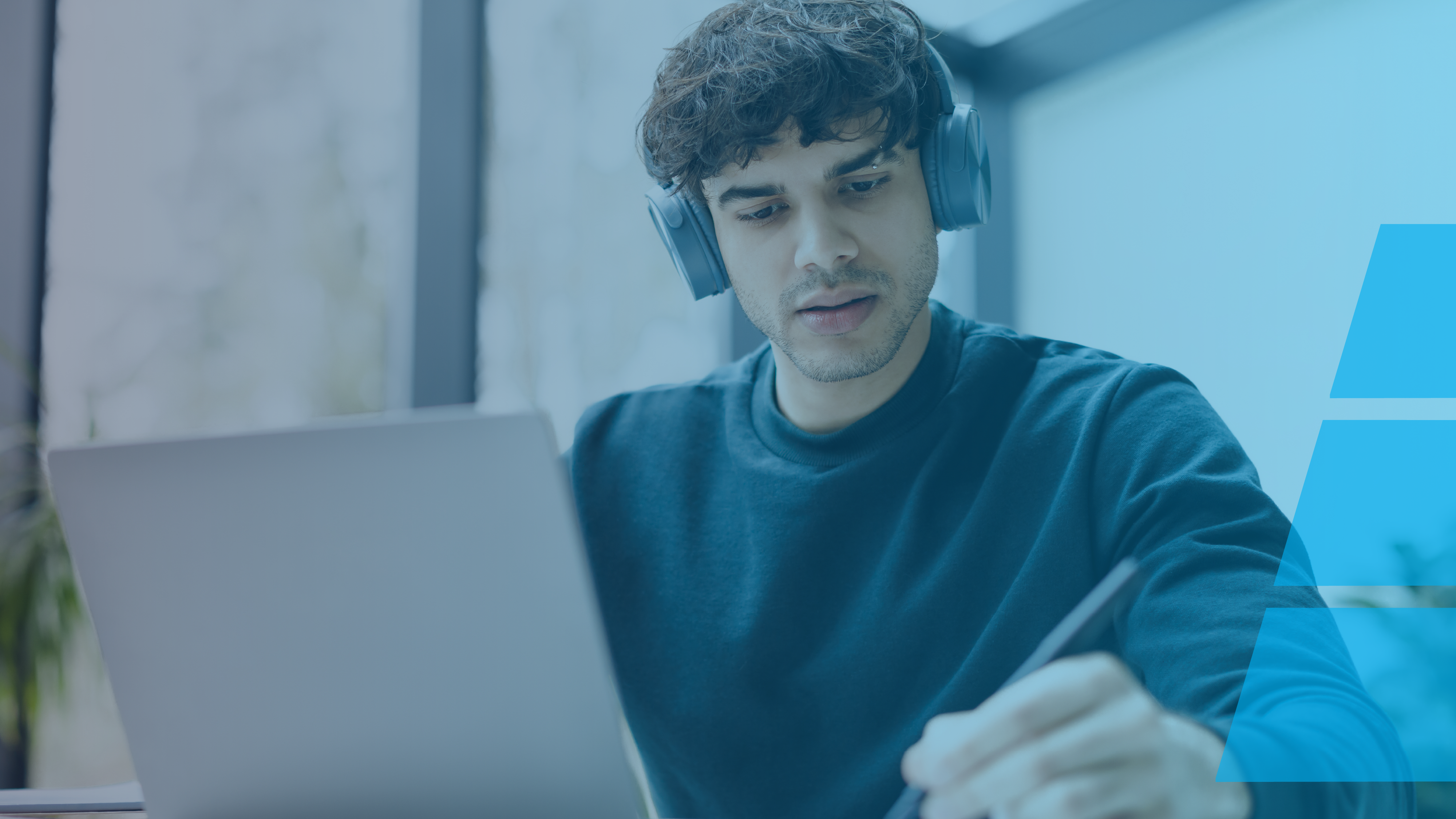 Young man wearing headphones and looking at a laptop while taking notes with a pen, seated indoors by a large window.